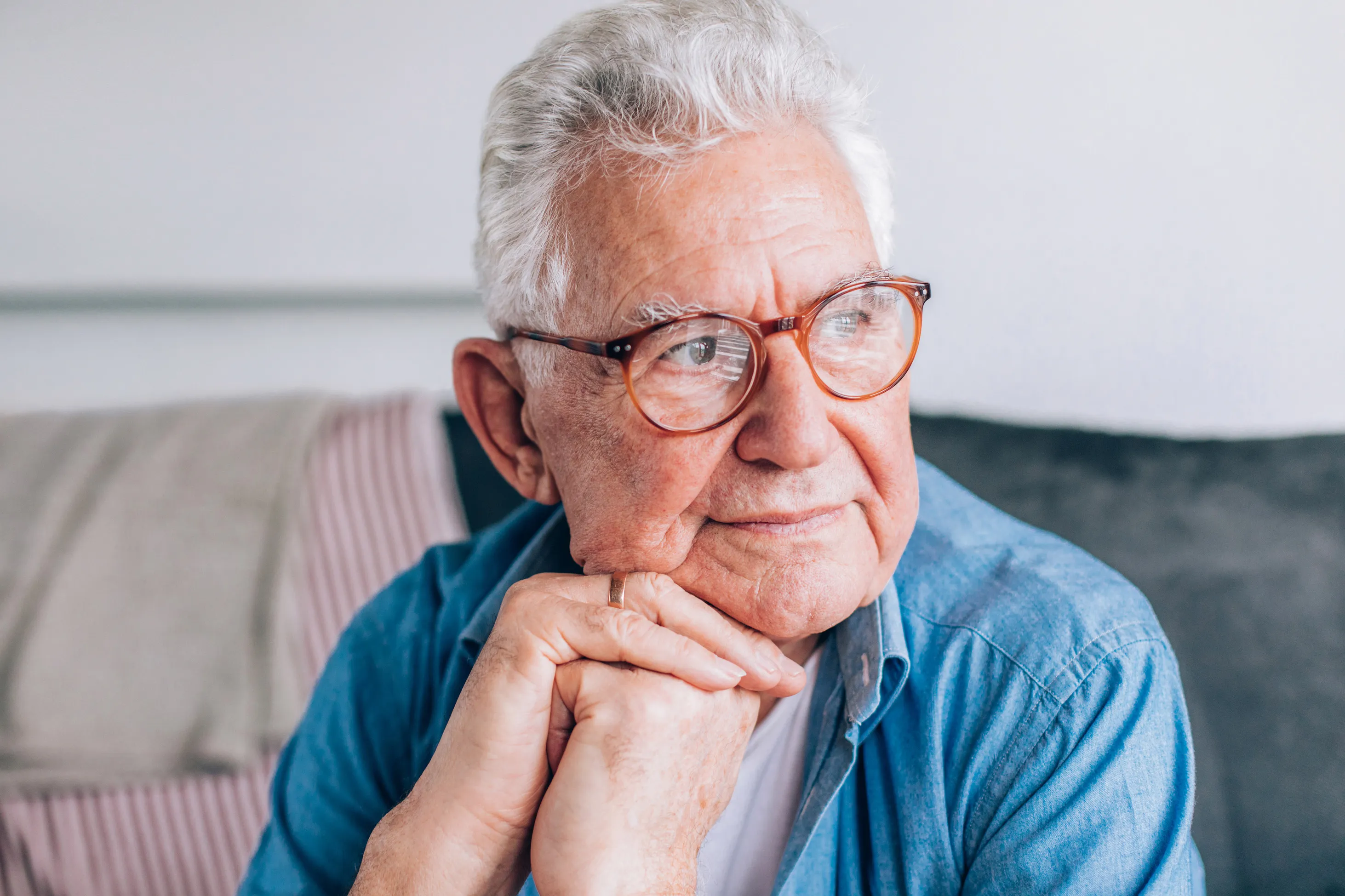 An older man in glasses sitting at home, resting his chin on his hands